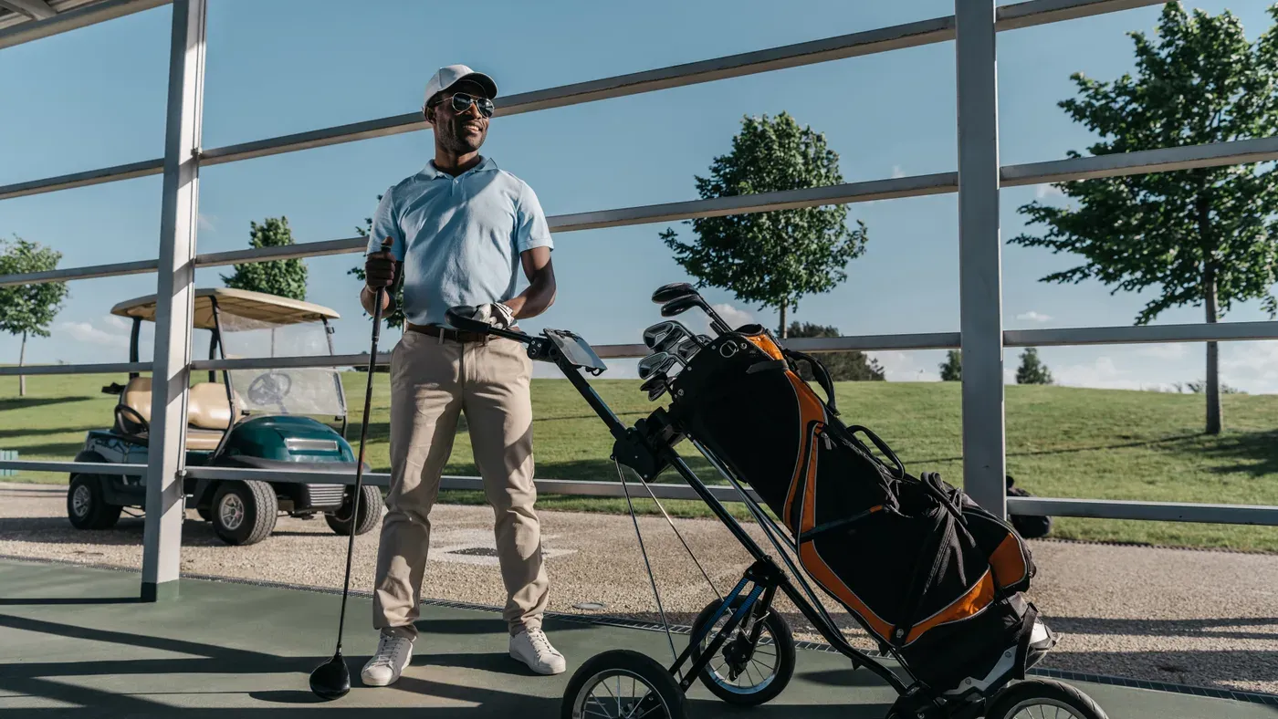 Golfare står med trehjulig vagn och fullpackad bag vid banan