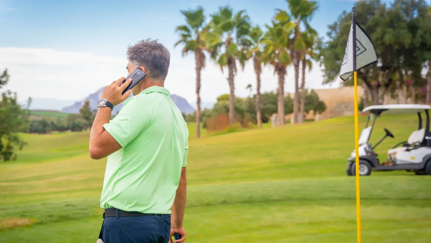 Golfare tittar på mobilen vid flaggan med golfbil i bakgrunden